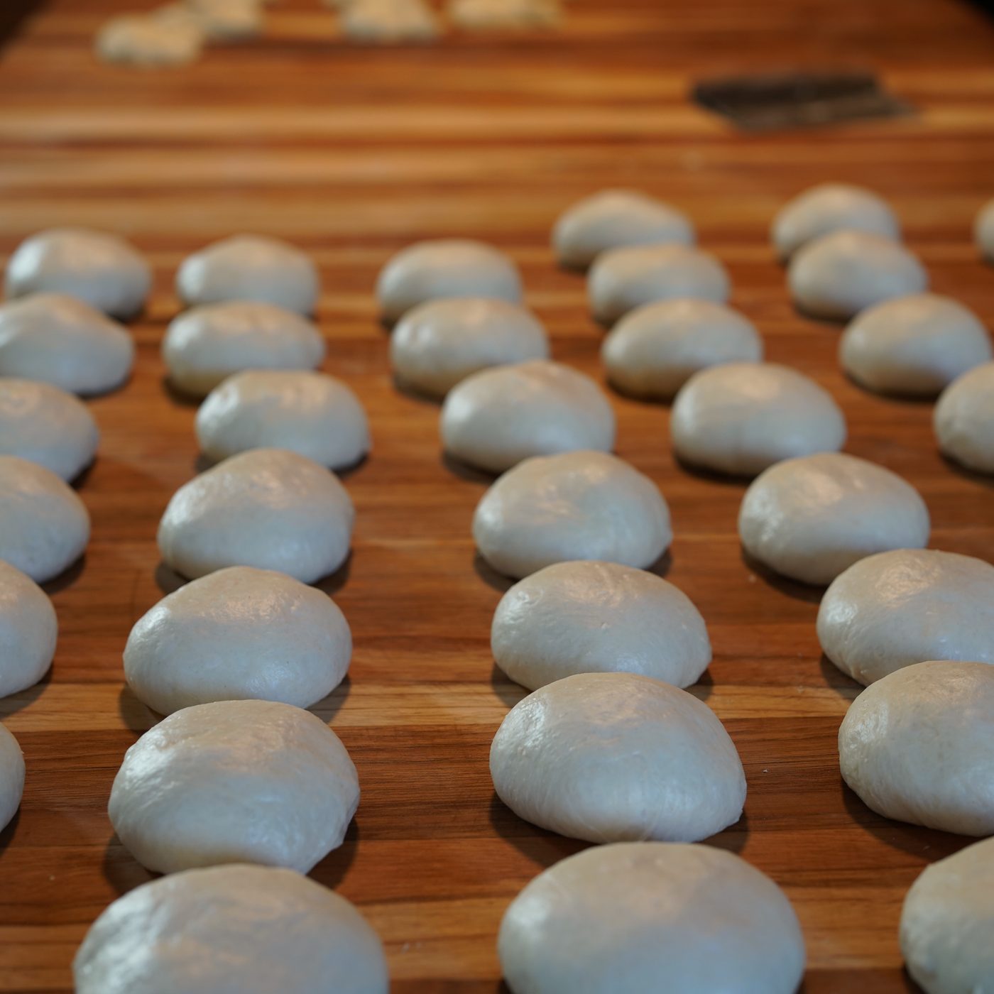Rows of smooth, pale dough balls resting on a striped wooden butcher-block bench, a bench scraper and more dough out of focus at the far end.