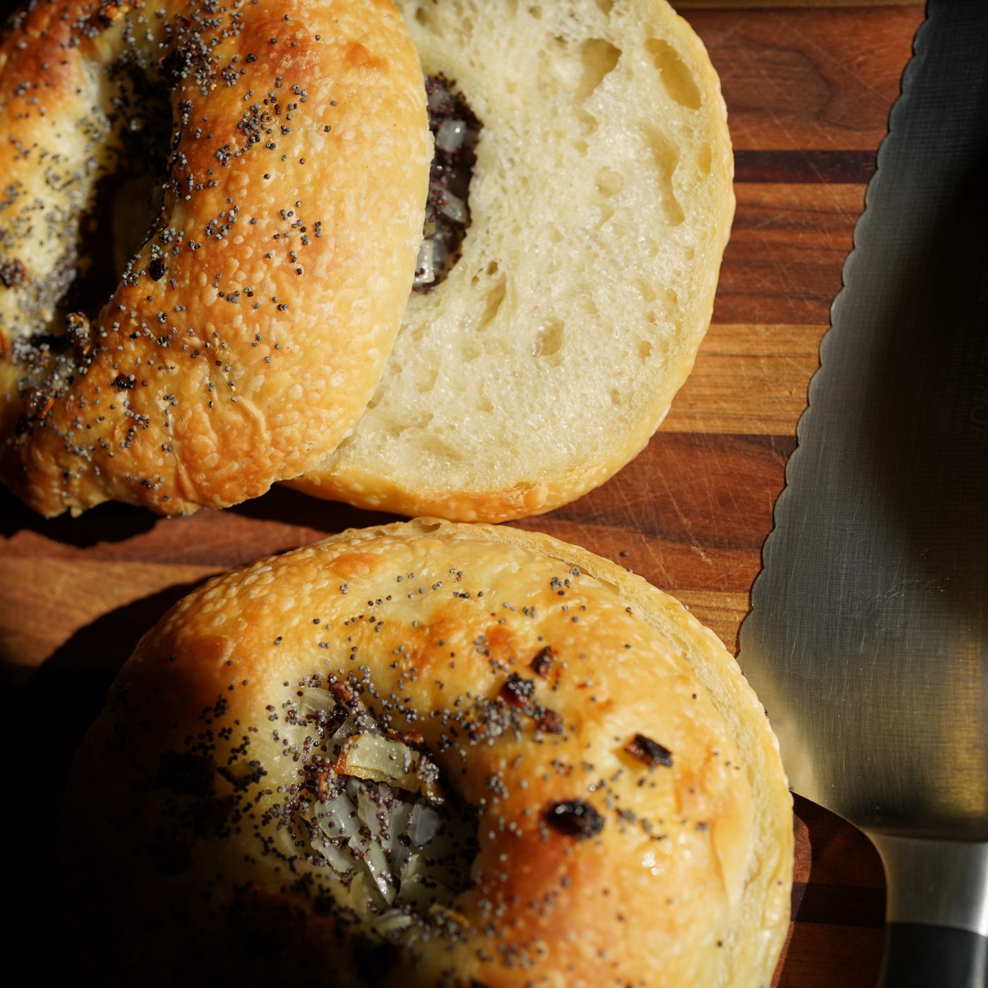 Two bialys on a wooden board beside a bread knife, one split open to show its crumb, the other whole with its center filled with onion and poppy seeds.