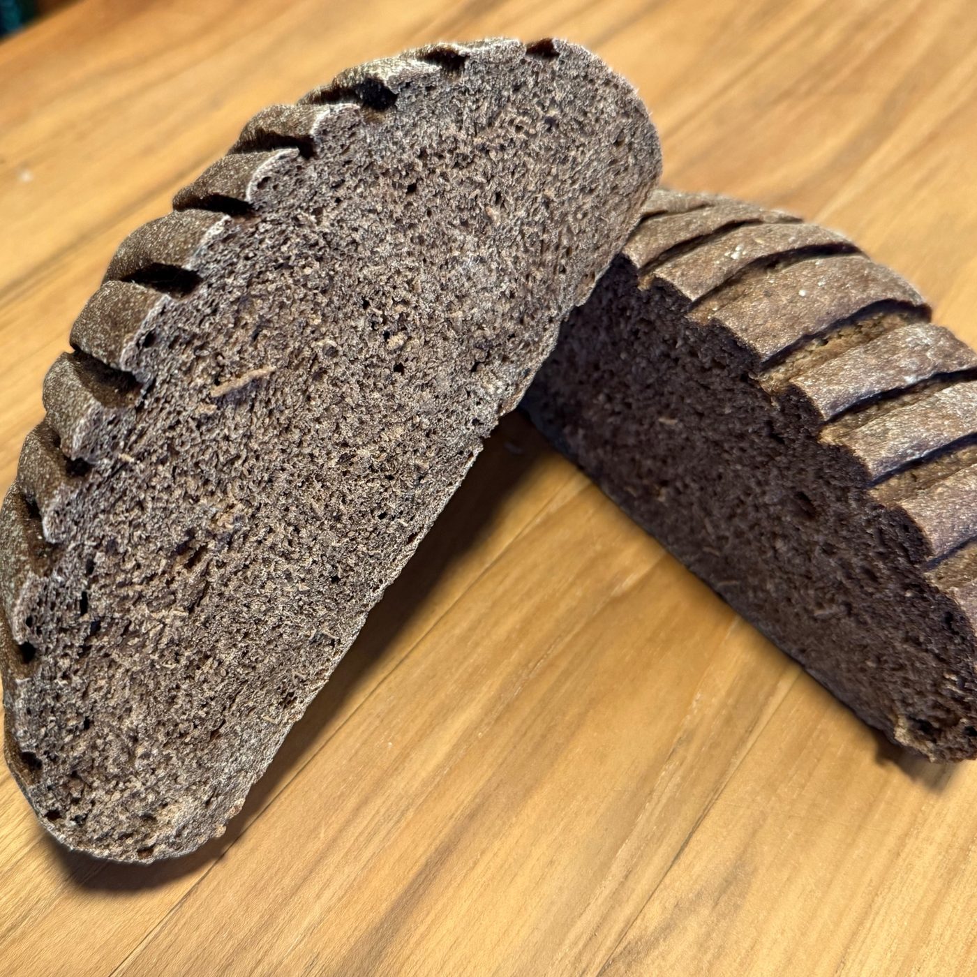 A round loaf of Serious Balt cut in half, the two halves leaning together on a wooden table to show a dense, dark brown crumb under a deeply scored crust.