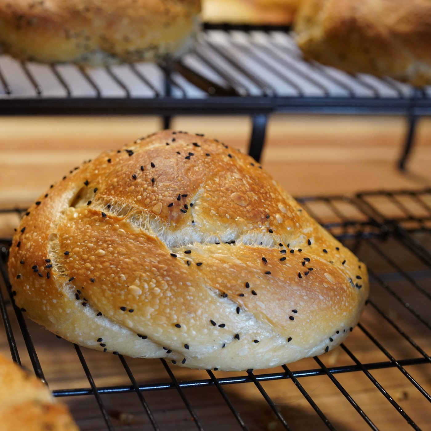 A Skally on a wire cooling rack, a golden braided loaf scattered with black nigella seeds, seeded rolls out of focus behind it.
