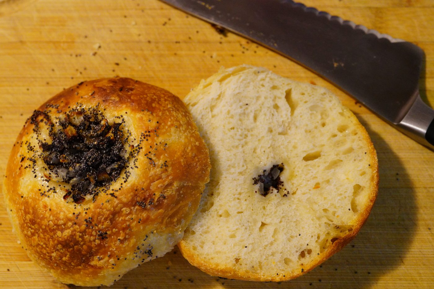 A bialy split in half on a wooden board, the filled centre of onion and poppy seeds on one half and the open crumb of the cut face on the other, a bread knife alongside.