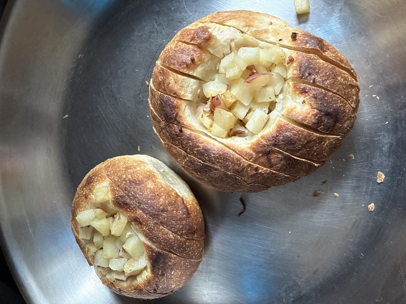 Two small round loaves in a steel pan, each scored across the top like a Hasselback potato, with diced potato and onion tucked into the cuts.