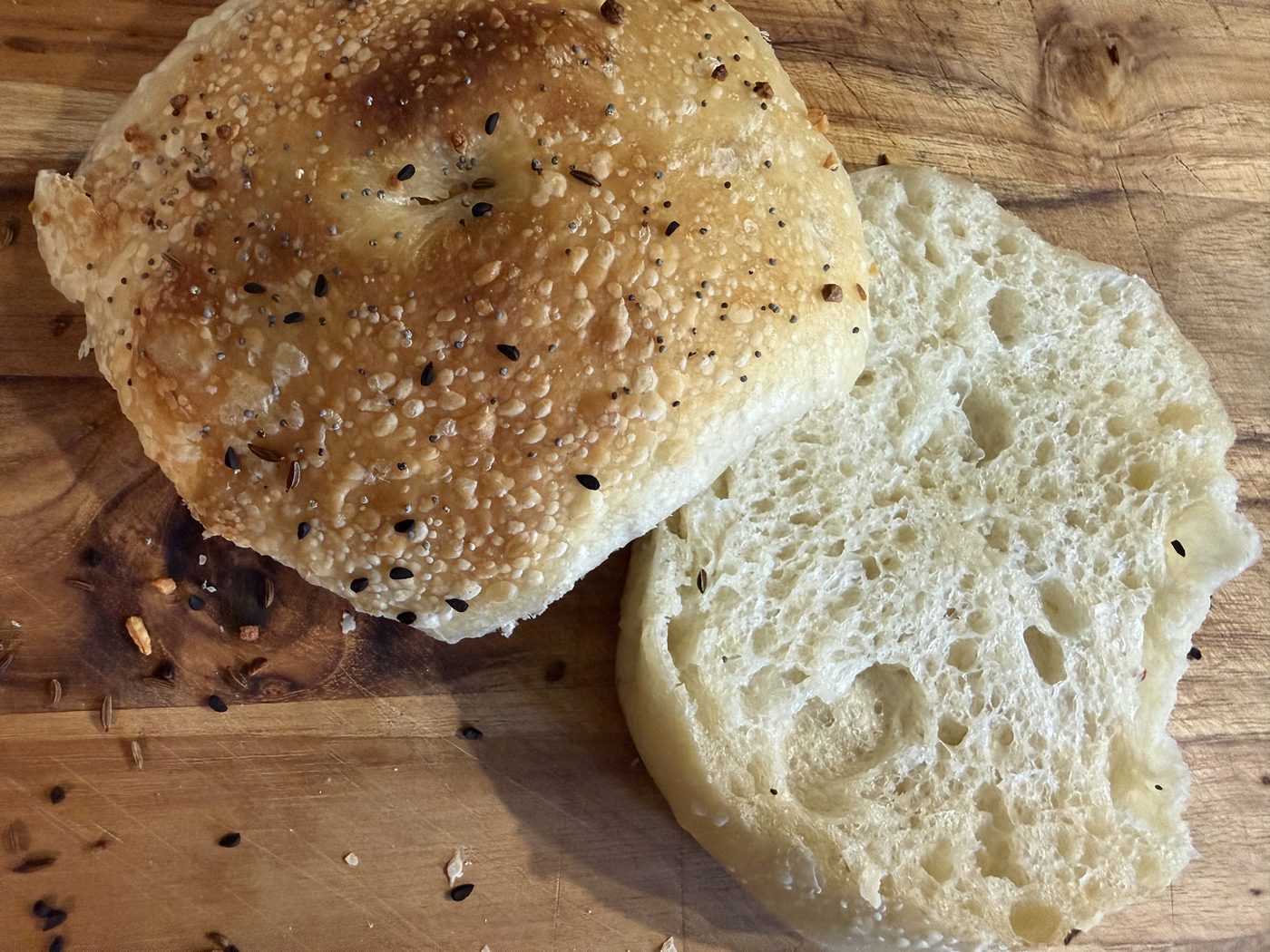 A split sourdough roll on a wooden board, its top crust blistered, golden and scattered with seeds, the cut half beside it showing a very open, airy crumb.